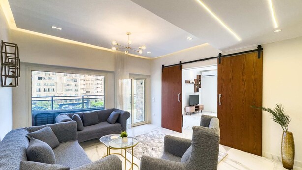 A modern living room with a grey sectional sofa, a glass coffee table, and a sliding door leading to another room.