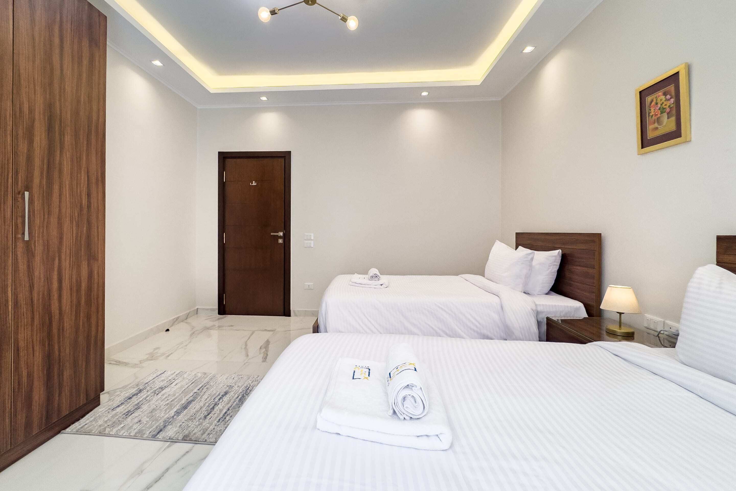 A hotel room with two beds, a wooden wardrobe, a marble floor, and a ceiling light fixture.