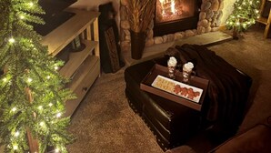 A cozy living room with a stone fireplace, a coffee table with cookies and a mug, and two Christmas trees with lights. - Mountain & Wildlife Views & Hot Tub Access (Estes Park)