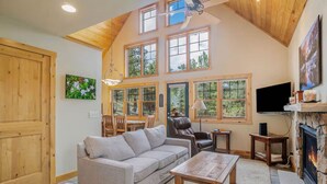 A cozy living room with a high ceiling, large windows, a fireplace, and a television. - Luxe Mountain Cabin! Views, jetted tub, deck, BBQ (Estes Park)