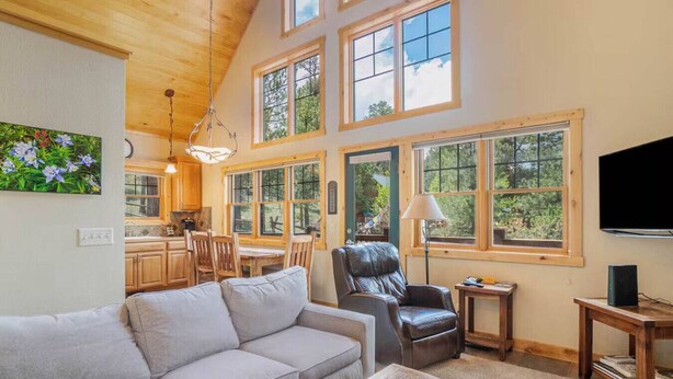 A spacious living room with a gray sofa, a leather chair, and a wooden coffee table. Large windows provide natural light and views of the outdoors. - Luxe Mountain Cabin! Views, jetted tub, deck, BBQ (Estes Park)