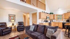 A spacious living area with a sofa, armchairs, and a coffee table. A kitchen with wooden cabinets and appliances is visible in the background. - Upstairs AC! Fireplace, deck, BBQ, shared hot tub (Estes Park)