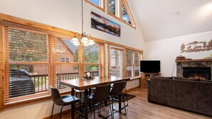 A spacious living area with a wooden dining table, chairs, a fireplace, and large windows with blinds. - Upstairs AC! Fireplace, deck, BBQ, shared hot tub (Estes Park)