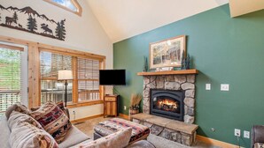A cozy living room with a stone fireplace, a television mounted above it, and a large window with blinds.