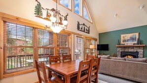 A dining area with a wooden table and chairs, large windows with blinds, a chandelier, and a fireplace in the living room.