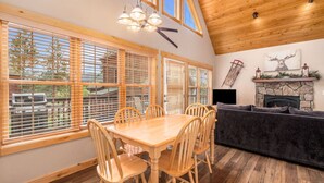 A dining area with a wooden table and chairs, large windows with blinds, a stone fireplace, and a cozy seating area with a sofa and fireplace mantel. - Best views! Deck, fireplace, BBQ, shared hot tub! (Estes Park)