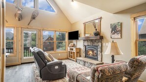 A cozy living room with a fireplace, leather sofa, patterned armchairs, and a large window overlooking a mountain. - Epic views, BBQ, deck, fireplace, shared hot tub! (Estes Park)