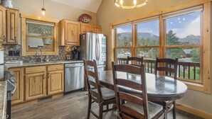 A kitchen with wooden cabinets, a stainless steel refrigerator, a dining table with chairs, and large windows offering a view of the outdoors. - Epic views, BBQ, deck, fireplace, shared hot tub! (Estes Park)