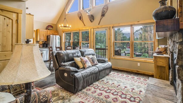 A spacious living room with a leather sofa, a patterned rug, and a large window offering a view of the outdoors. - Epic views, BBQ, deck, fireplace, shared hot tub! (Estes Park)