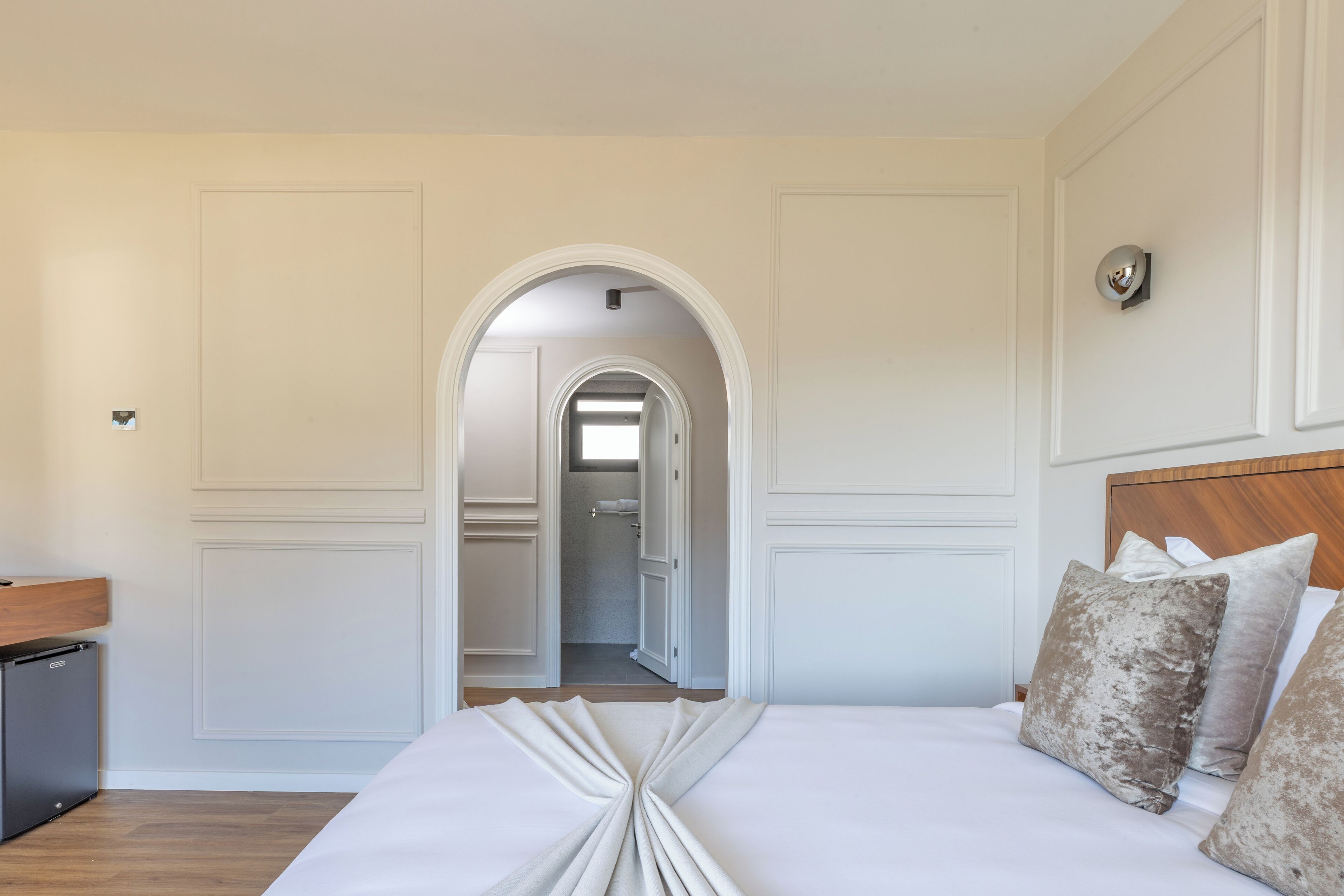 Habitación de hotel con una cama bien hecha, cabecera de madera y vista al baño a través de un arco.