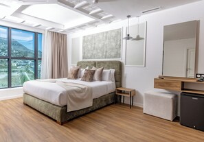 A modern bedroom with a large bed, a nightstand, a small refrigerator, and a view of a mountainous landscape through large windows. - Aurea Hotel (Tetouan)
