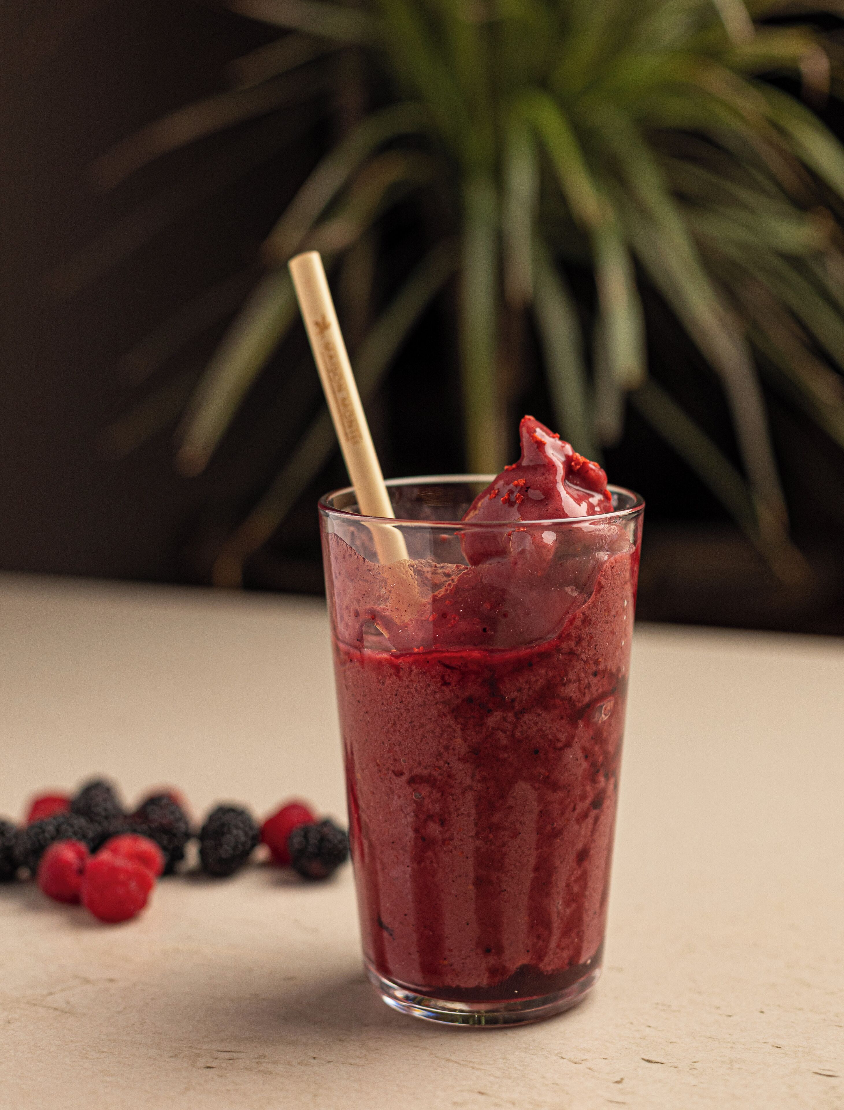 A glass of red smoothie with a straw, surrounded by berries on a table.