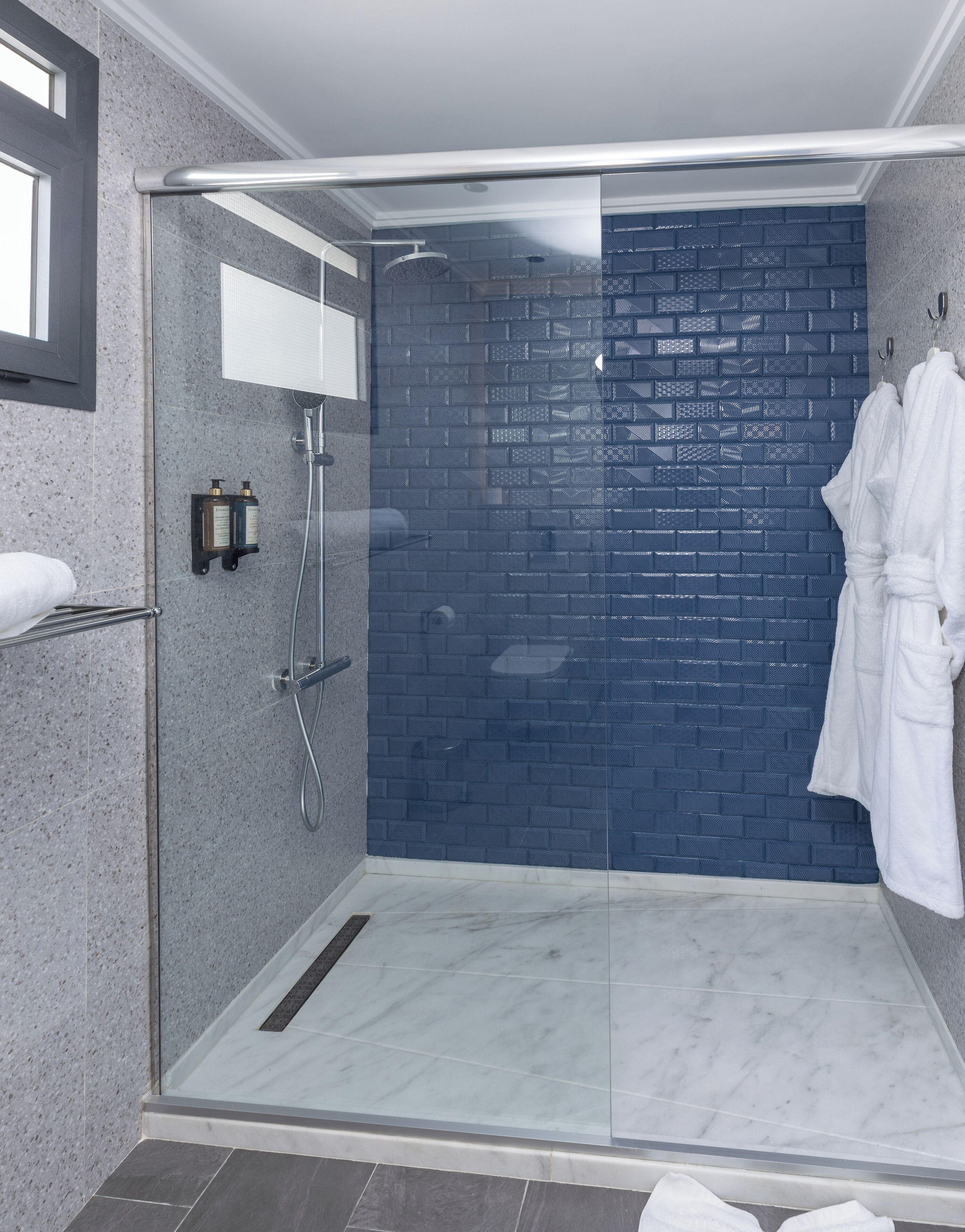 Un área de ducha moderna con puerta de cristal, pared de ladrillo azul y una bata blanca colgada en la pared.