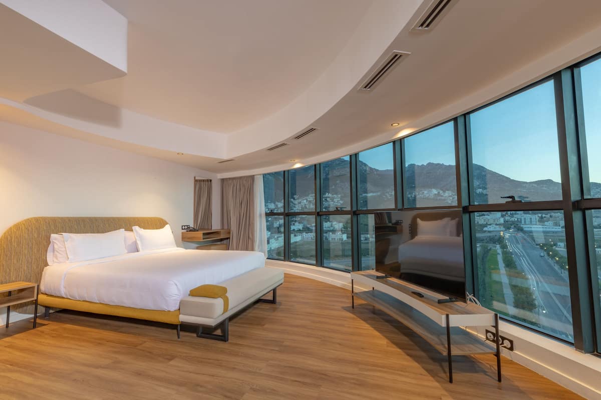 Una habitación de hotel moderna con una cama grande, televisión y vistas a un paisaje urbano a través de amplios ventanales.