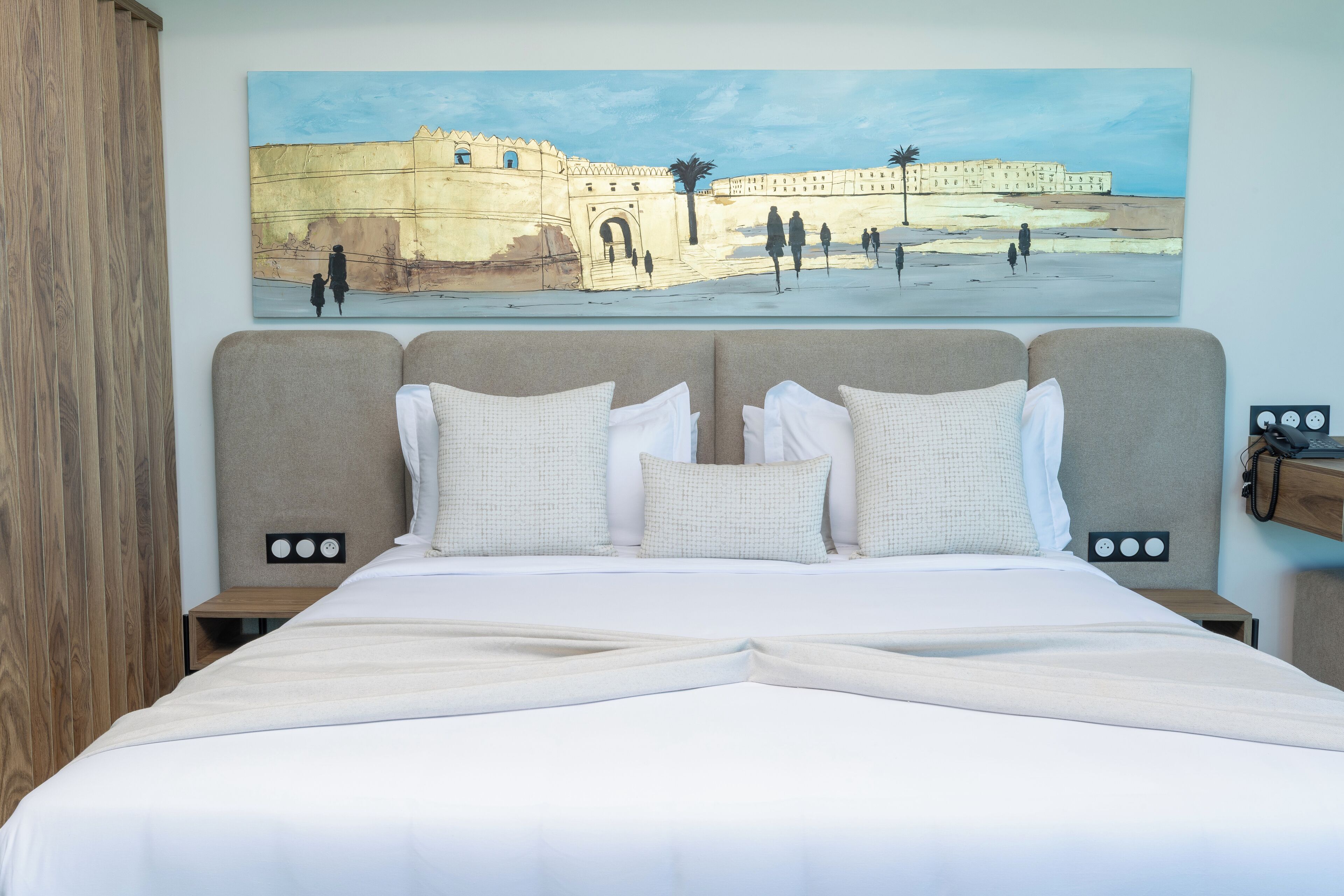 A hotel room with a large bed, two bedside tables, and a painting of a historical building on the wall.