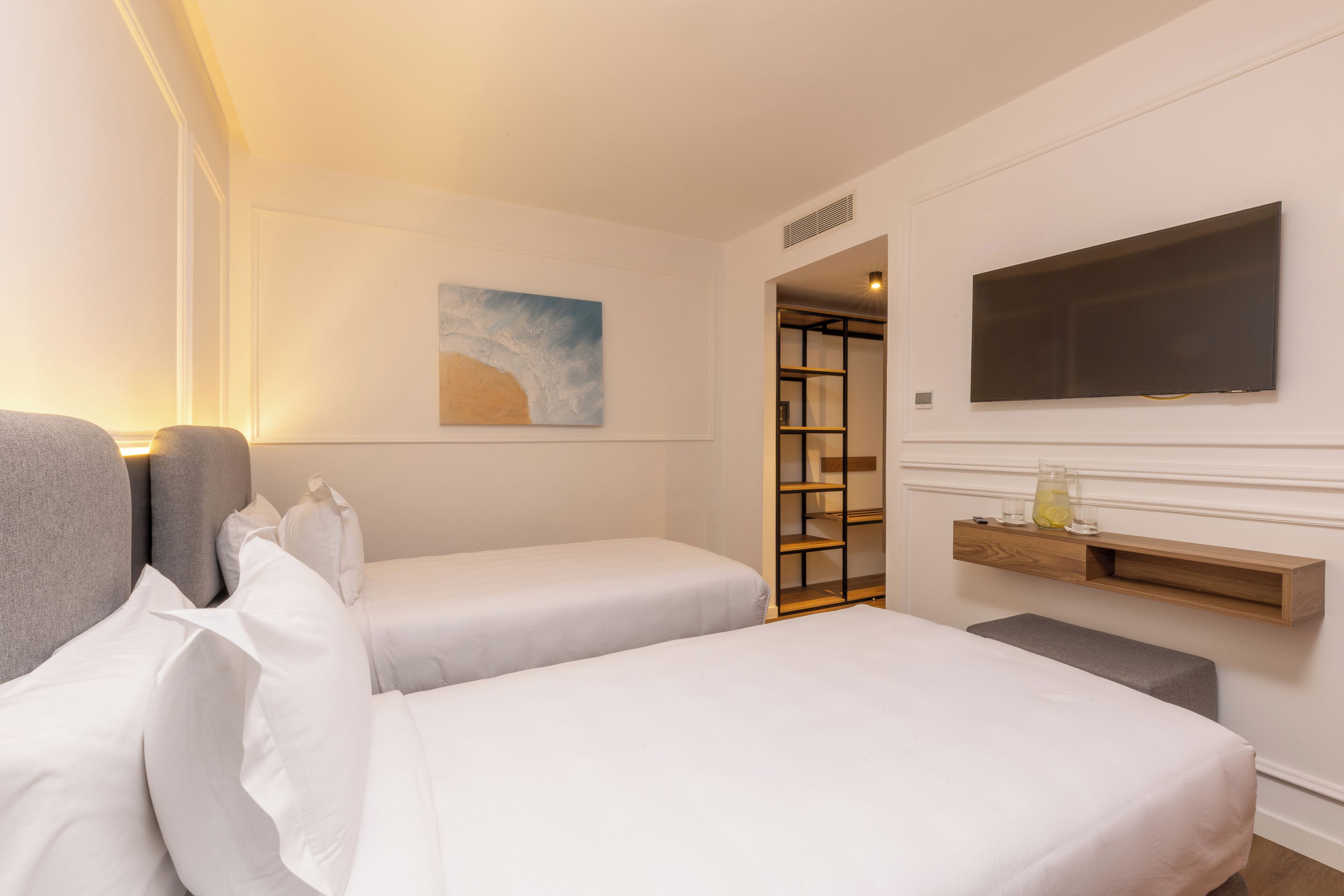 Habitación de hotel con dos camas individuales, un televisor de pantalla plana montado en la pared, una mesita de noche con lámpara y un cuadro en la pared.