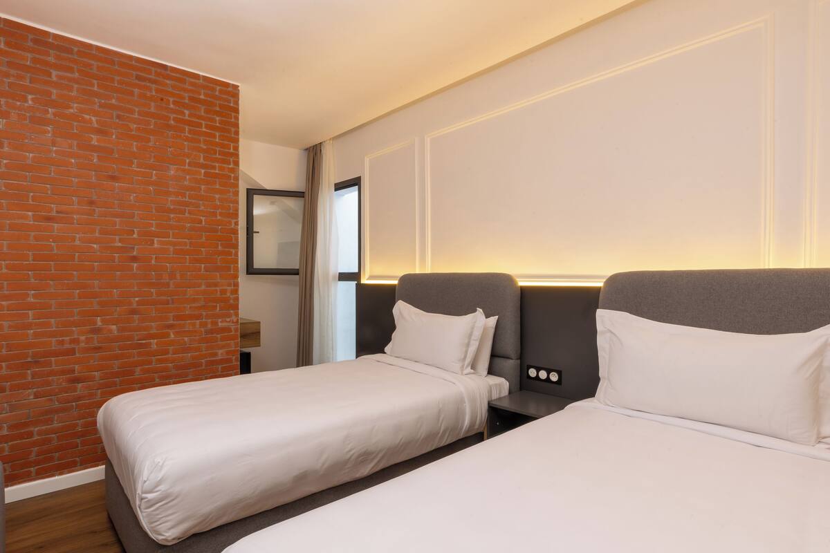 Habitación de hotel con dos camas individuales, una pared de ladrillo, una ventana con cortinas y un panel de control en la pared.