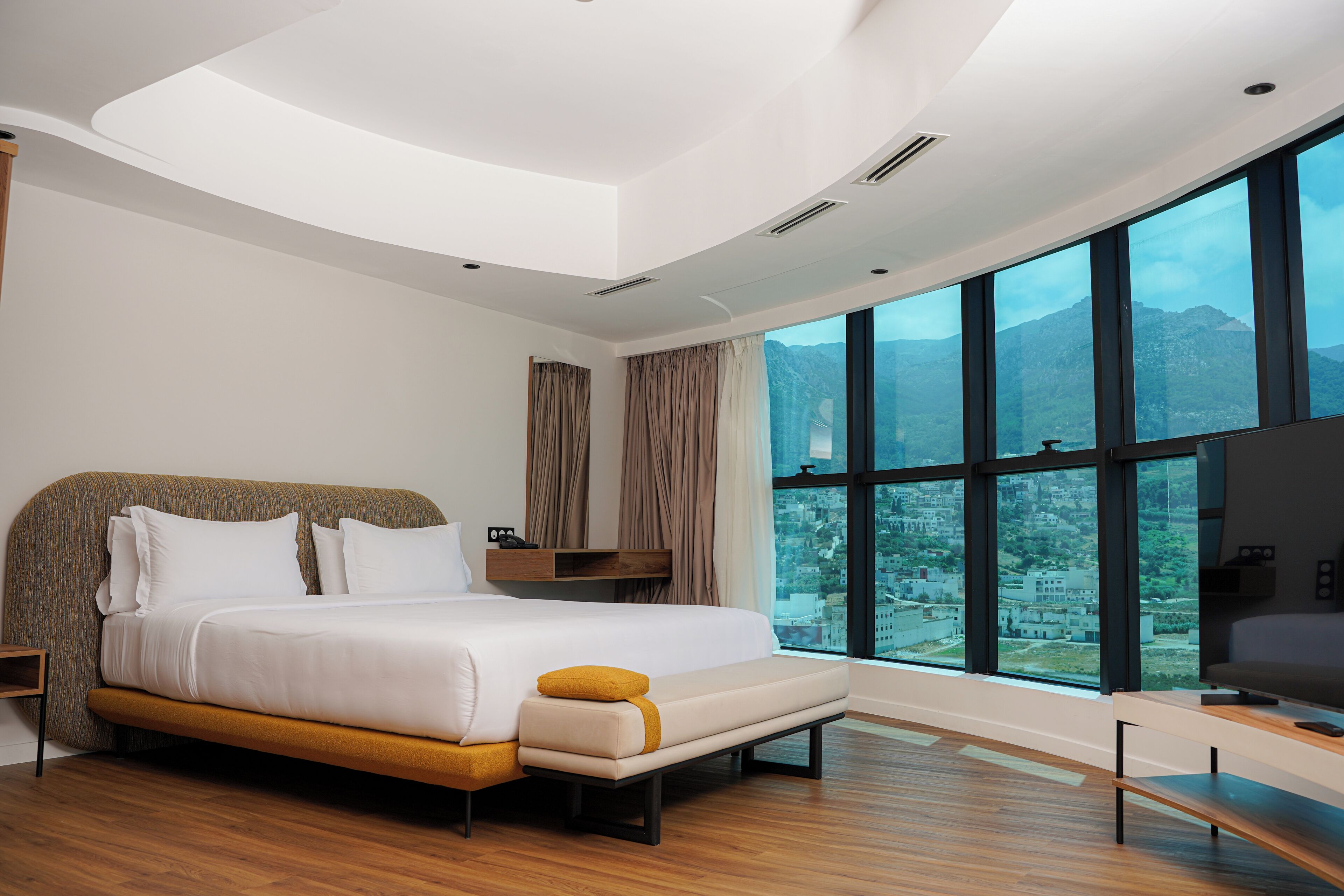 Un dormitorio moderno con una cama grande, un banco, un televisor y vistas a montañas a través de amplios ventanales.