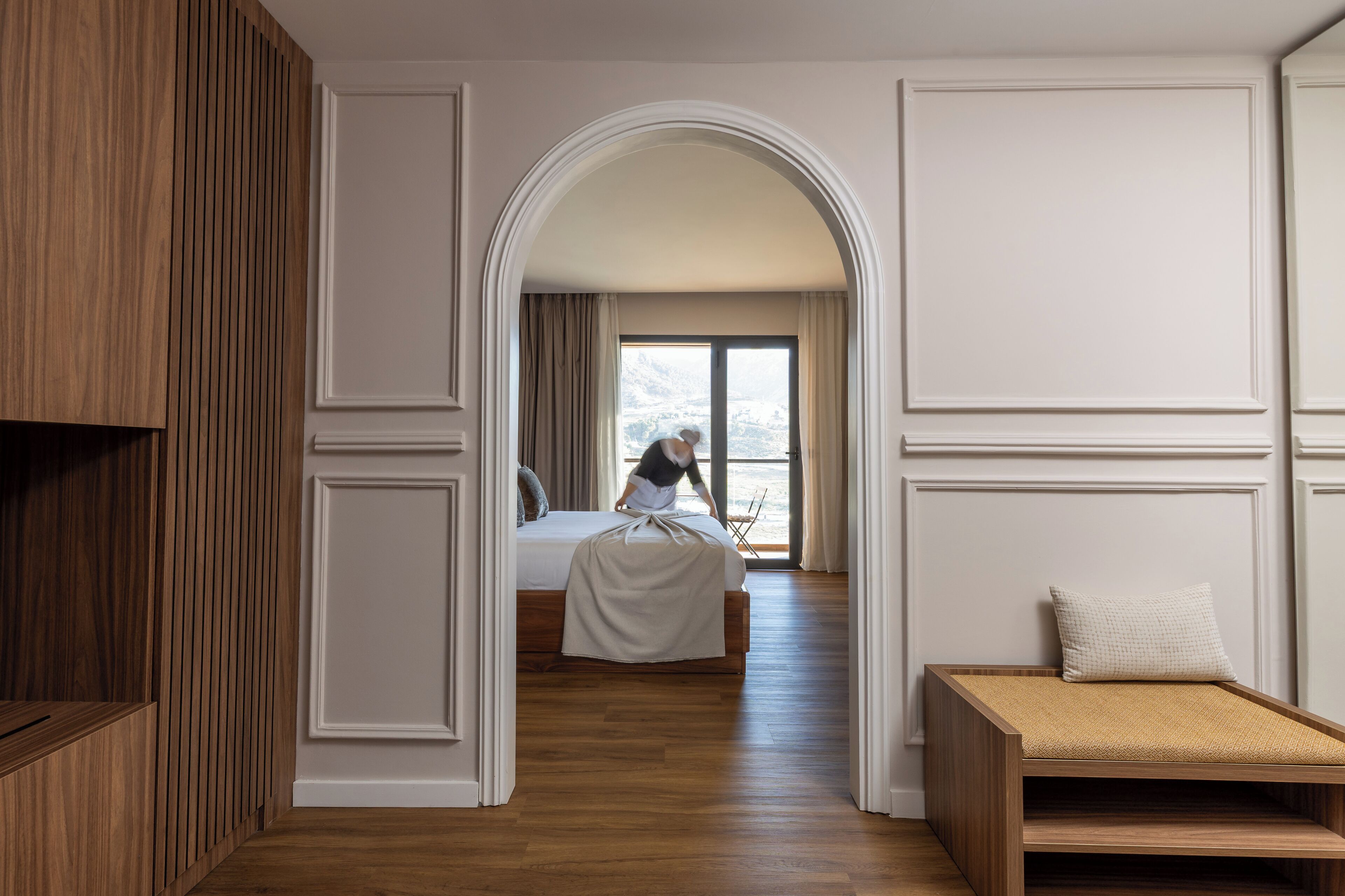Une chambre d’hôtel avec un plancher en bois, un lit recouvert d’une courtepointe blanche, un banc en bois et une vue sur les montagnes par une grande fenêtre.