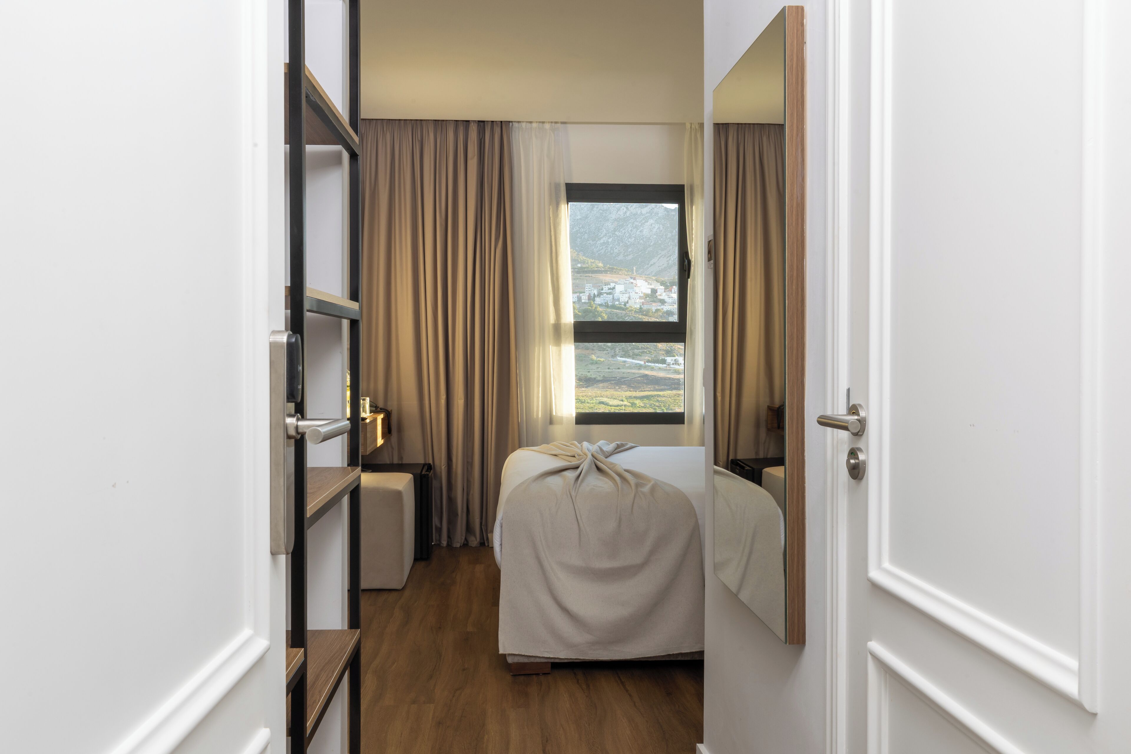 Habitación de hotel con cama, mesita de noche, cortinas y vistas a un paisaje montañoso.