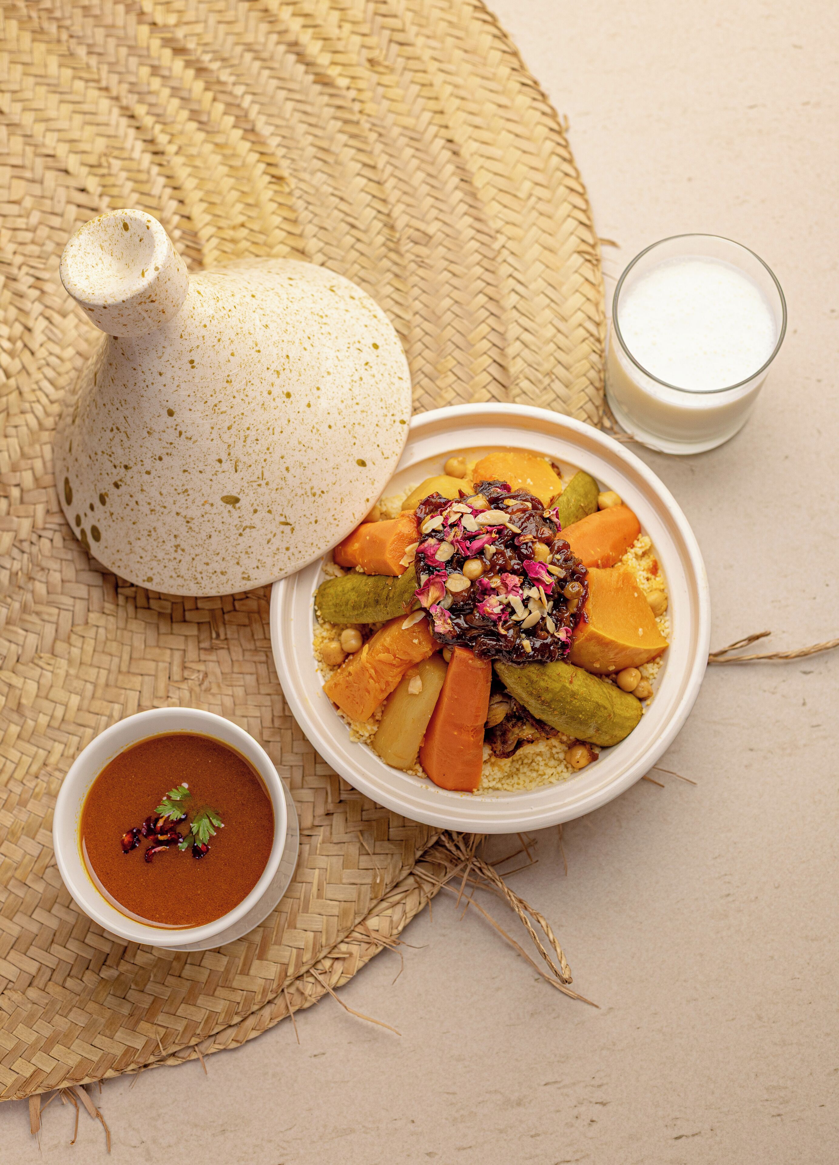 A meal with couscous, vegetables, and a sauce, served on a woven mat with a teapot and a glass of milk.