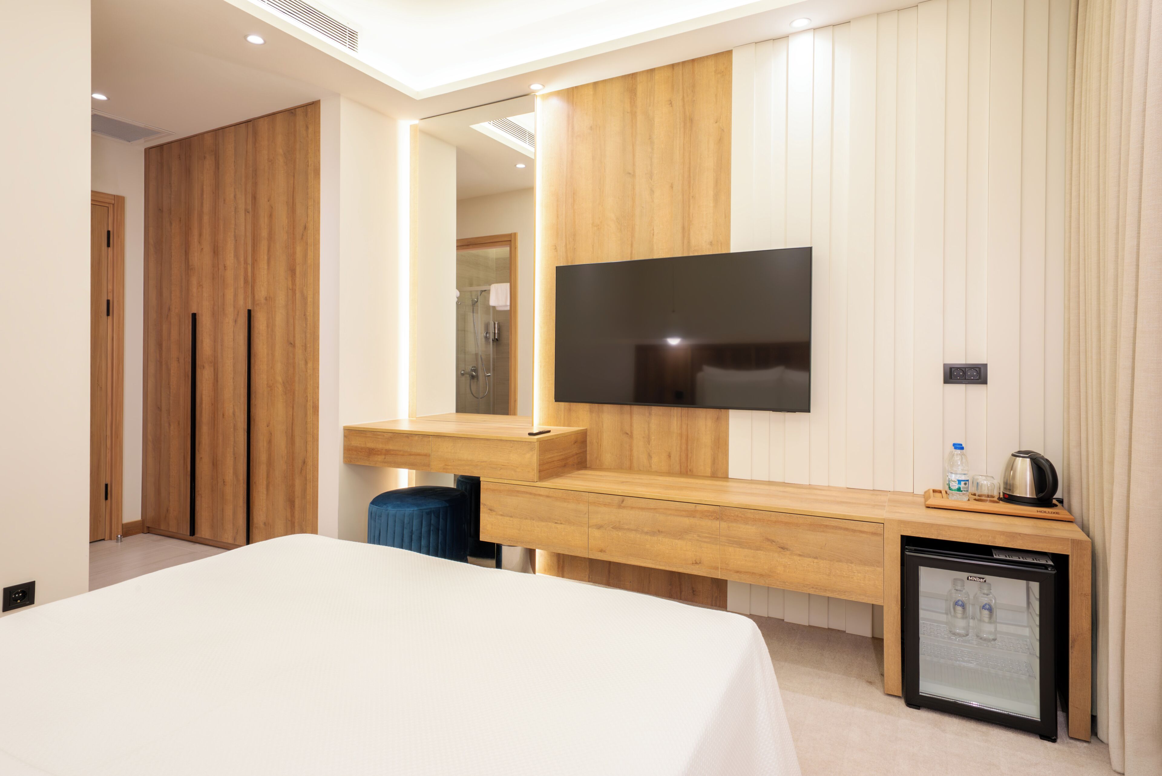 A modern hotel room with a bed, a wooden vanity, a television, a minibar, and a small refrigerator.