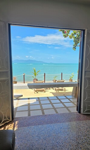 Une porte ouverte donne sur une terrasse offrant une vue sur la mer, agrémentée de plantes en pot et de mobilier d’extérieur.