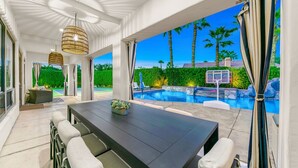 A modern outdoor dining area with a pool view, featuring a long table, cushioned chairs, and a hanging light fixture. - 0.4 Acre- Indio Sapphire Oasis with Game Room (Indio)