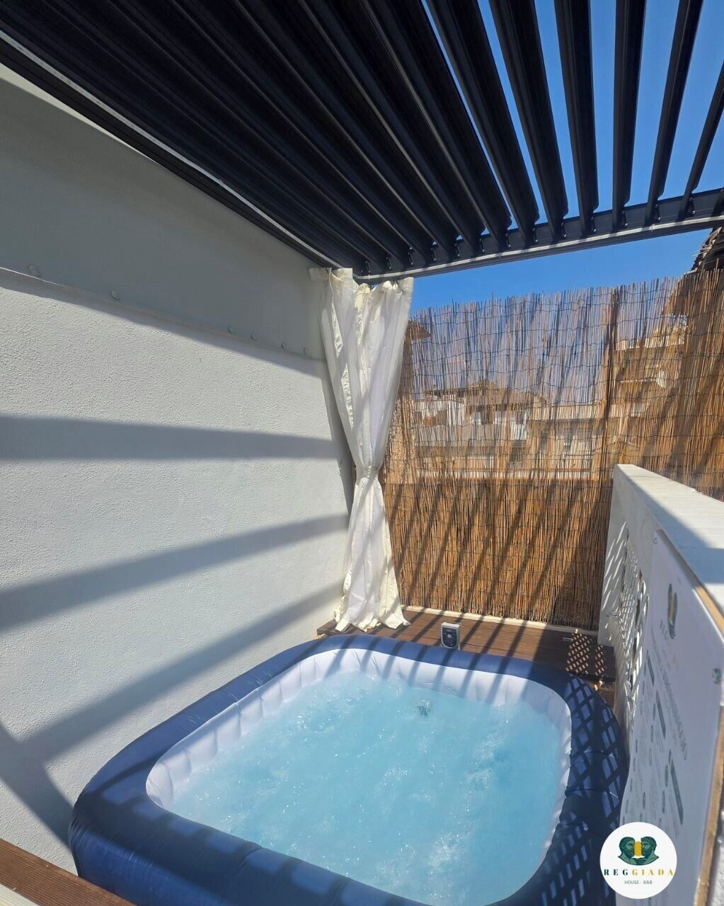 An outdoor hot tub area with a pergola overhead, a wooden deck, and a white wall.