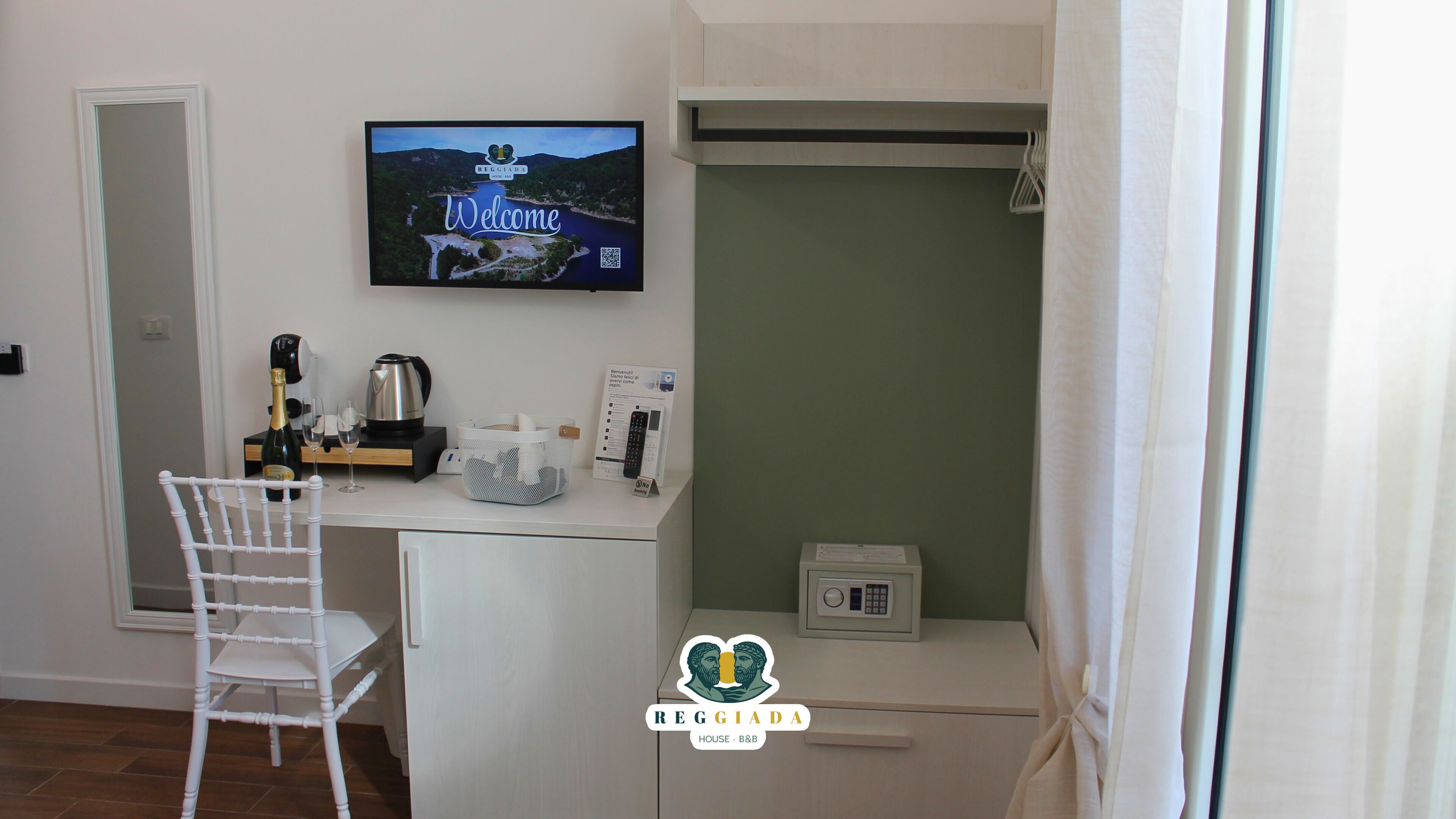 A small kitchenette with a TV displaying a welcome message, a white chair, a bottle of champagne, and a coffee maker.