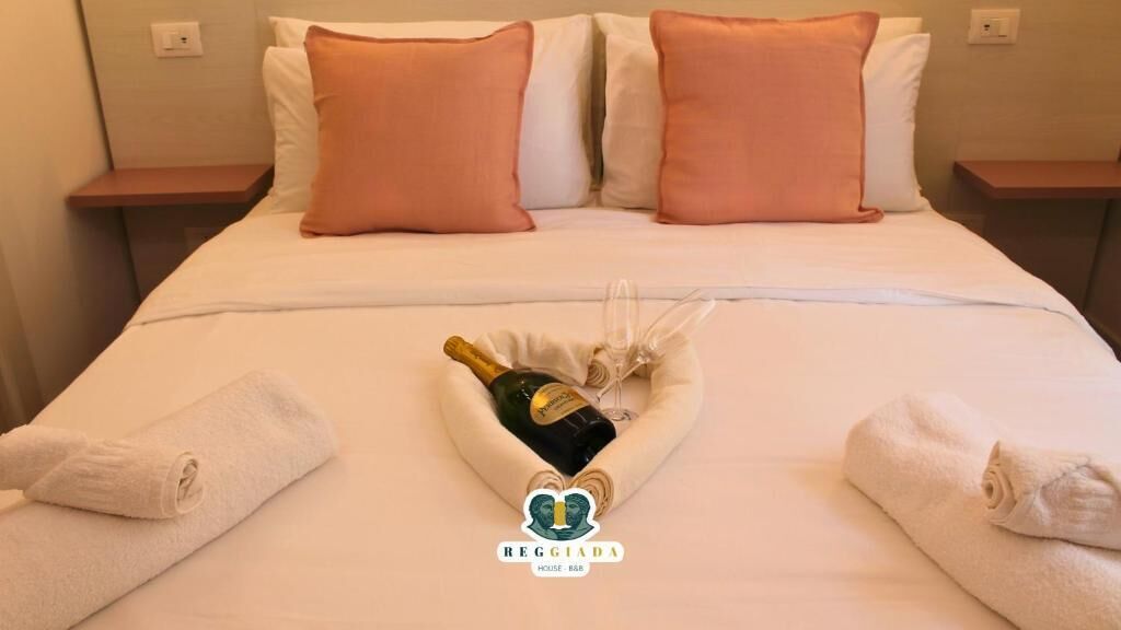 A hotel room with a neatly made bed, two pillows, a bottle of champagne, and a glass.