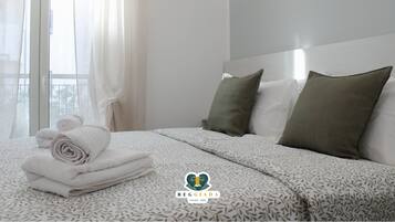 Una camera d'albergo con un letto rifatto, una finestra con tende leggere e un logo sul copripiumino.