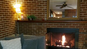 A living room with a brick fireplace, a sofa with a white cushion, a wall-mounted lamp, and a ceiling fan.