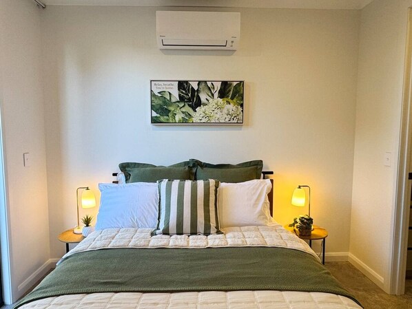 A bedroom with a bed, two bedside lamps, a wall-mounted air conditioner, and a framed picture above the bed.