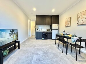A modern living room with a flat-screen TV, a dining table with chairs, a kitchen area with dark cabinets, and a microwave.