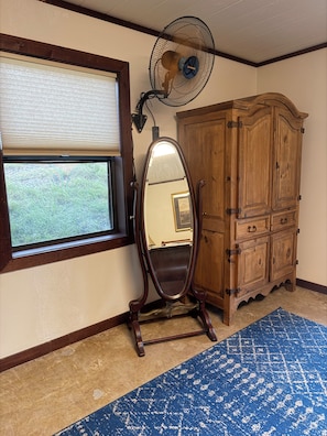Habitación con ventilador de pie, un espejo grande, un armario de madera y una ventana con persianas.