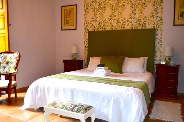 A neatly made bed with a green headboard, a chair with a floral pattern, and a small bedside table with a lamp.