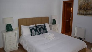 A neatly made bed with white linens, two decorative pillows, and a folded towel. There are two bedside tables with lamps and a radiator on the wall. - This is a double room with a double bed. It has an en-suite bathroom. The window in the room looks out into the hallway. (Vilaflor)