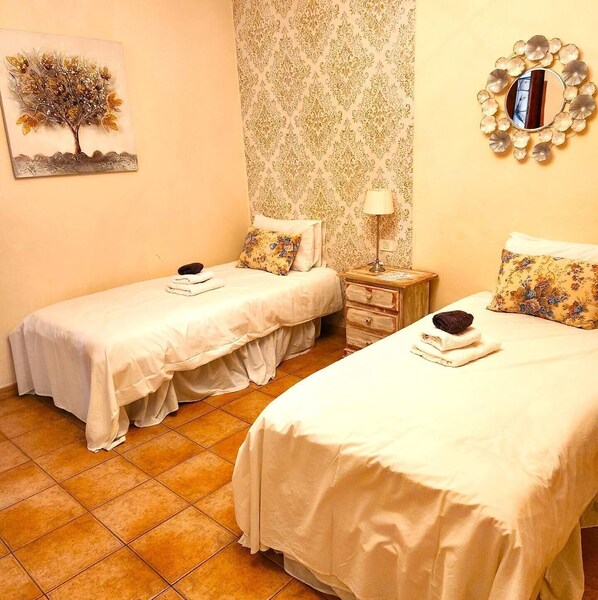 Una habitación con dos camas, cada una con una colcha blanca y una almohada. Hay un espejo adosado a la pared con un marco decorativo, una mesita con una lámpara y un cuadro de un árbol en la pared.