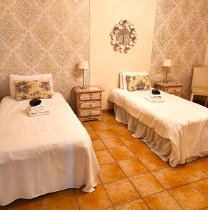 Una habitación con dos camas, cada una con un cojín floral y una lámpara. Hay un espejo en la pared y una silla en la esquina.