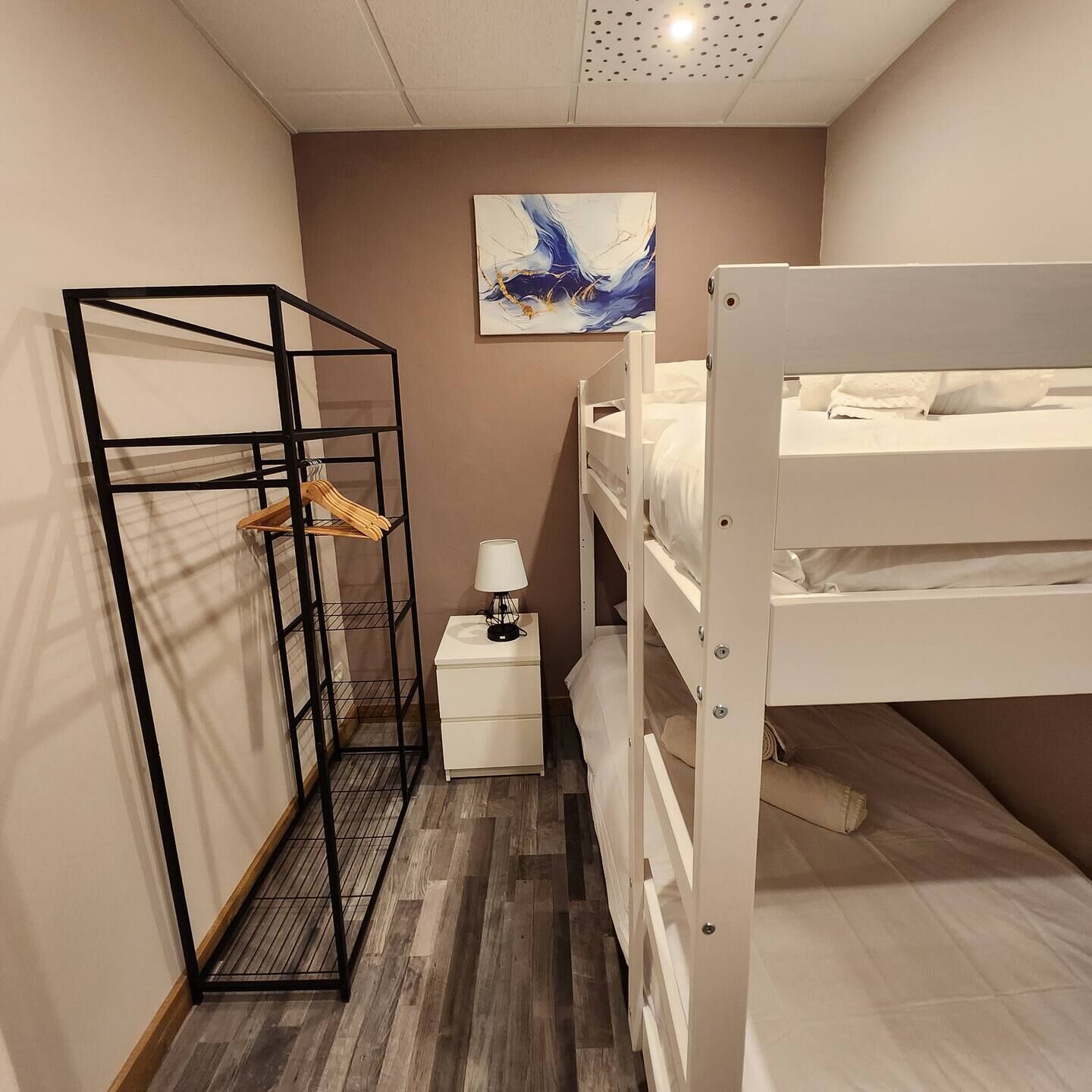 A compact room with a bunk bed, a small bedside table with a lamp, a hanging rack, and a painting on the wall.
