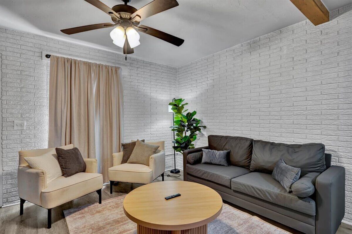 A living room with a white brick wall, a ceiling fan, a beige sofa, a grey sofa, two armchairs, a coffee table, and a floor lamp.