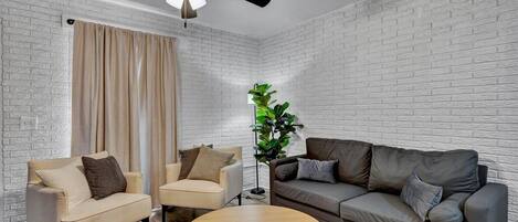 A living room with a white brick wall, a ceiling fan, a beige sofa, a grey sofa, two armchairs, a coffee table, and a floor lamp.