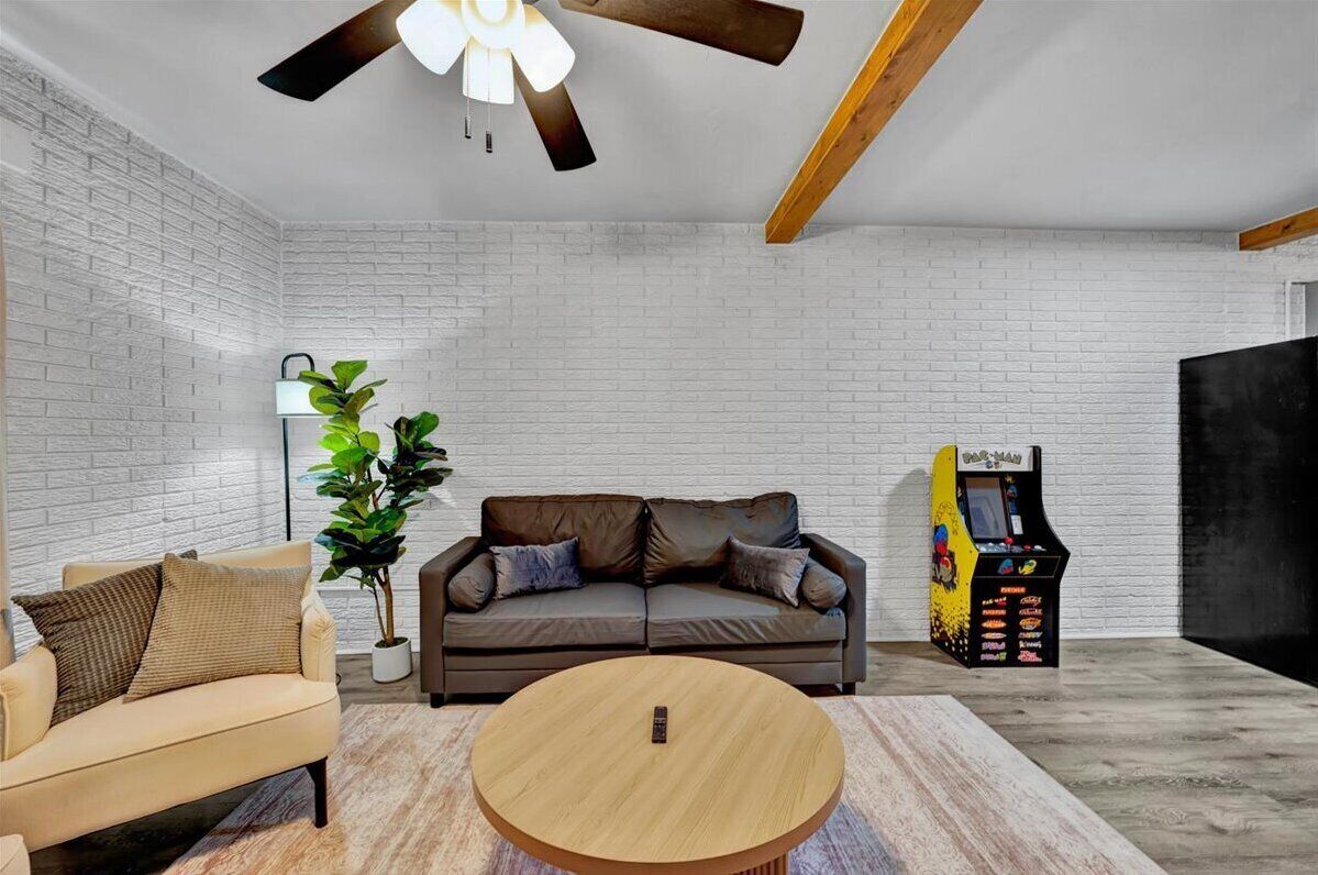 Una sala de estar moderna con una pared de ladrillo blanco, un sofá de cuero marrón, un sillón beige, una mesa de centro de madera y una máquina de arcade retro.