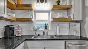 A modern kitchen with white cabinetry, a black countertop, and wooden beams. There is a window above the sink, and various kitchen items are visible. - Family-Friendly Stay Near OSU & Boomer Lake (Stillwater)