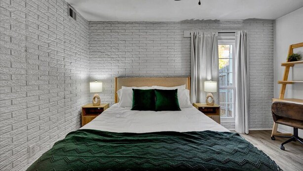A bedroom with a brick-patterned wall, a wooden headboard, a bed with white linens and a green throw, two bedside tables with lamps, and a window with curtains. - Family-Friendly Stay Near OSU & Boomer Lake (Stillwater)