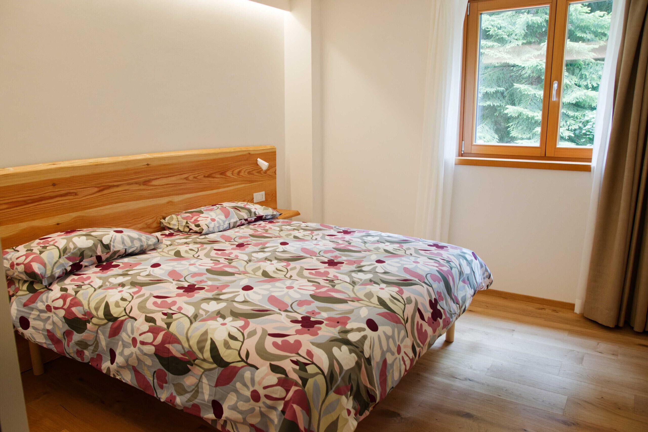 Un dormitorio con cabecera de madera, colcha estampada con motivos florales y ventana con vista a árboles.