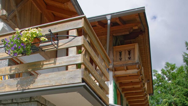 Une cabane en bois à deux étages, avec des murs en pierre, un balcon orné de jardinières suspendues et un chemin de pierre menant à l’entrée.