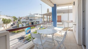 Un balcon avec une table et des chaises blanches, donnant sur une rue bordée de boutiques et un ciel dégagé.