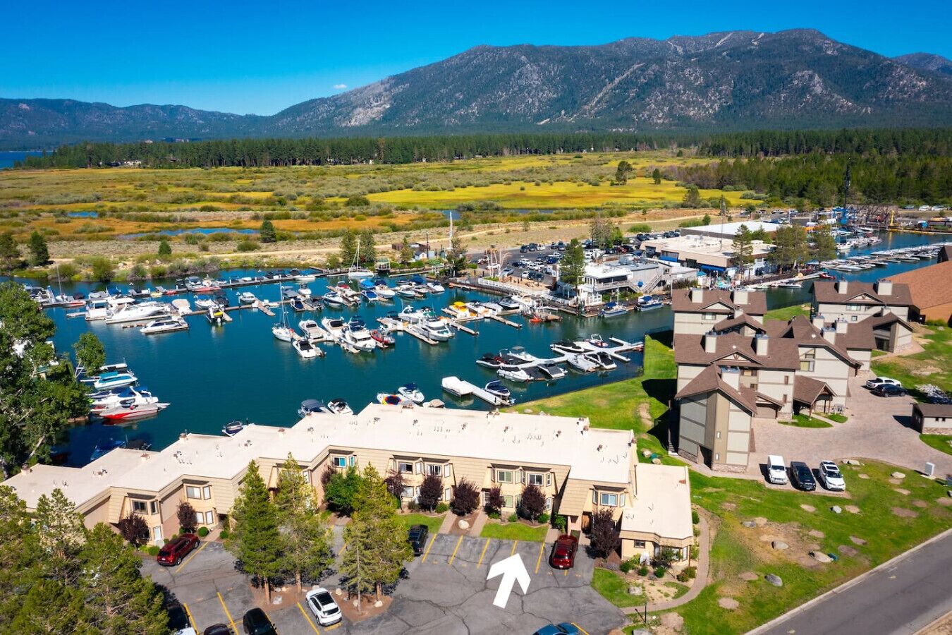 A marina with boats, a building with a parking lot, and mountains in the background.